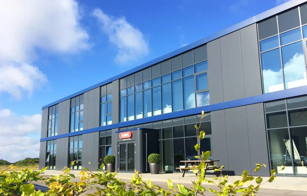 Bildet viser en moderne kontorbygning med store glassvinduer og en mørk fasade med blå detaljer. Foran inngangen står to store krukker med planter og et piknikbord, omgitt av grønne busker under en klarblå himmel.