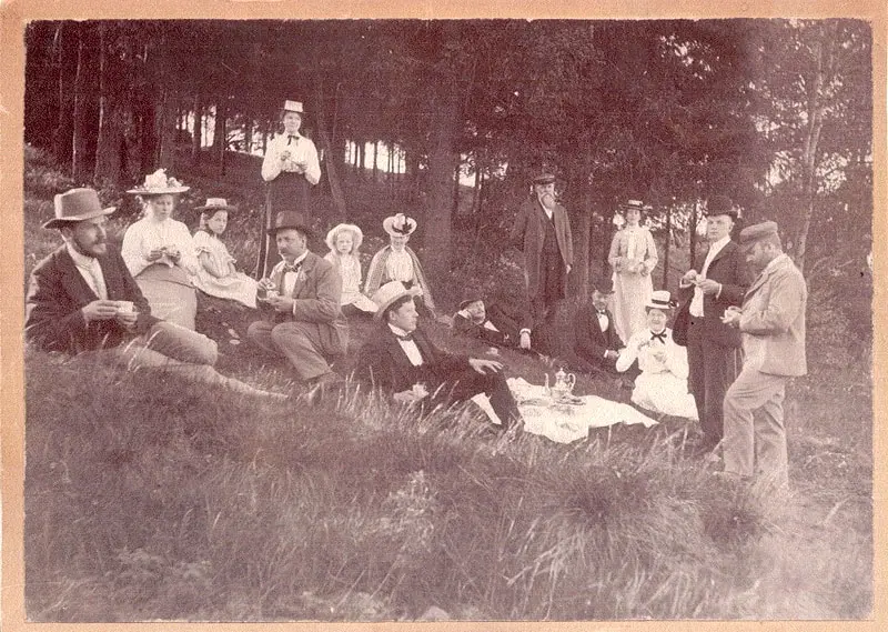 Et veldig gammelt og gulnet bilde der velkledde herrer og damer i hatt, samt to barn, har piknik på en eng.