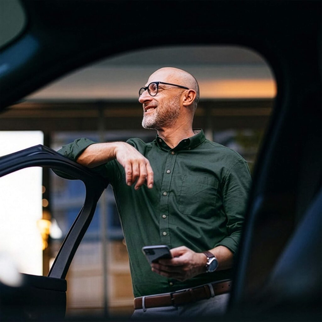 En person står med mobilen i hånden ved en bil og ser utover.